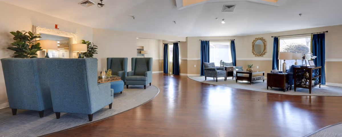 A spacious senior living facility common area with wooden flooring and two separate seating areas. The left seating area has blue high-back armchairs and a small table with decorative items. The right seating area features armchairs, a coffee table, side tables with lamps, and large windows with blue curtains letting in natural light. The walls are painted beige with white trim, and there are plants and a mirror adding to the decor.