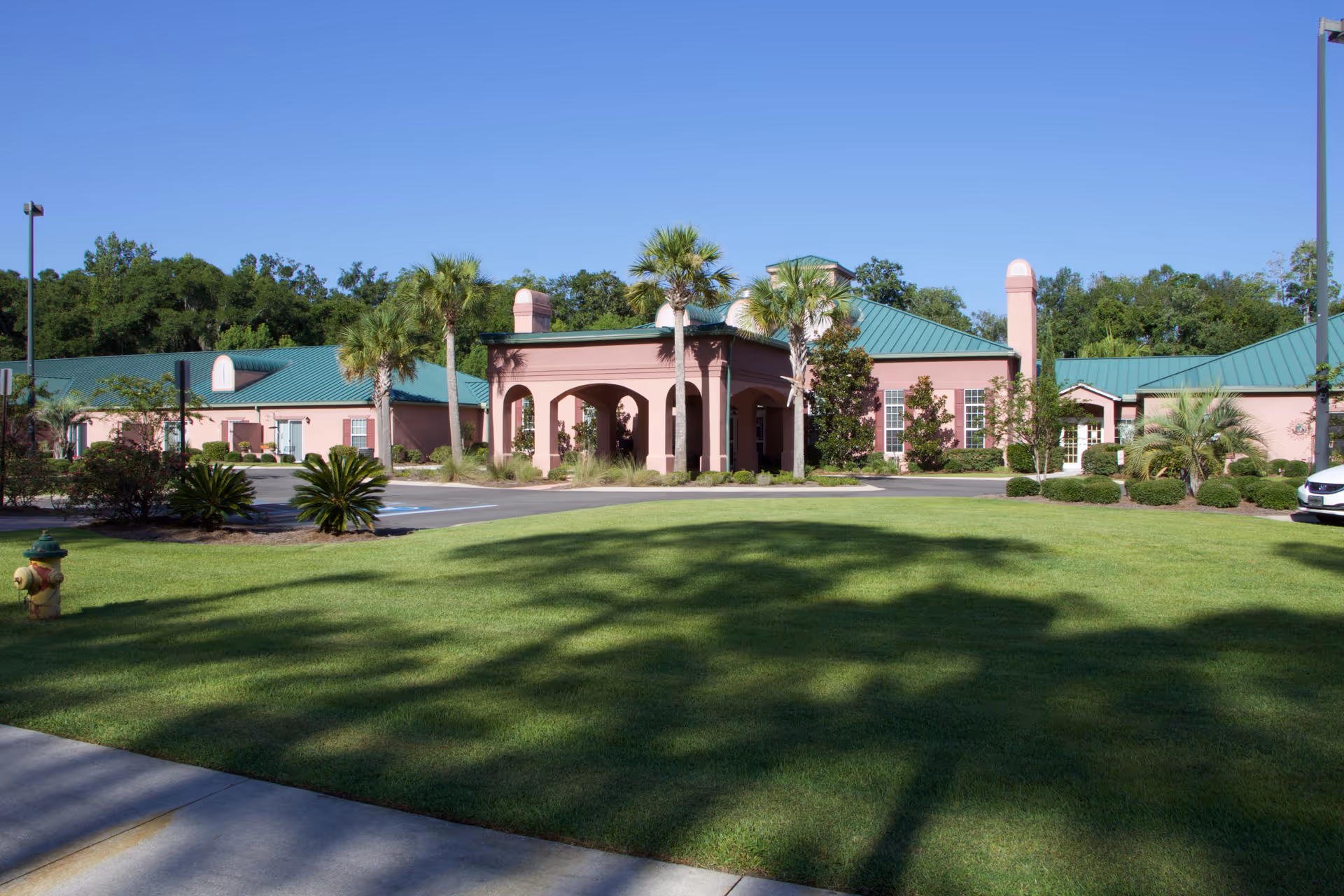 Front exterior of a single-story pink senior living facility with a covered entrance, green metal roof, palm trees and a manicured lawn.
