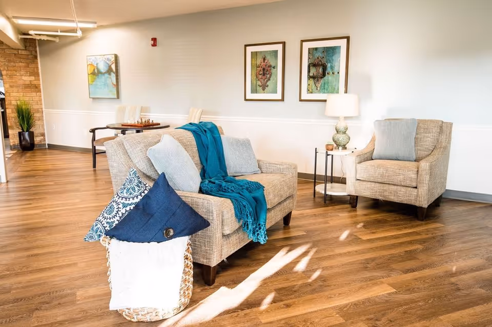A cozy seating area in a senior living facility with a beige loveseat and armchair, both adorned with light blue cushions. A teal throw blanket is draped over the loveseat. There is a small round side table with a decorative lamp and small plants. The room has wooden flooring, light-colored walls with two framed artworks, and a basket filled with pillows in the foreground. In the background, there is a small table with a chessboard and chairs, and a potted plant near a brick wall.