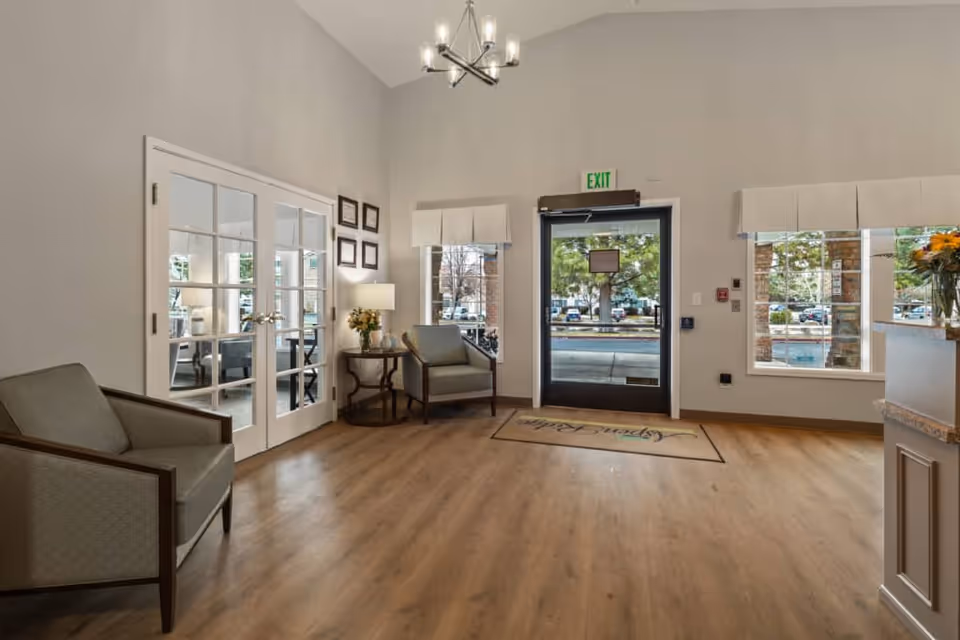 A bright and welcoming lobby area with wooden flooring, two armchairs, a small round table with a lamp and flowers, large windows with white valances, a glass door with an exit sign above it, and a chandelier hanging from the ceiling. There is a doormat with the text 'Aspen Ridge' near the entrance.