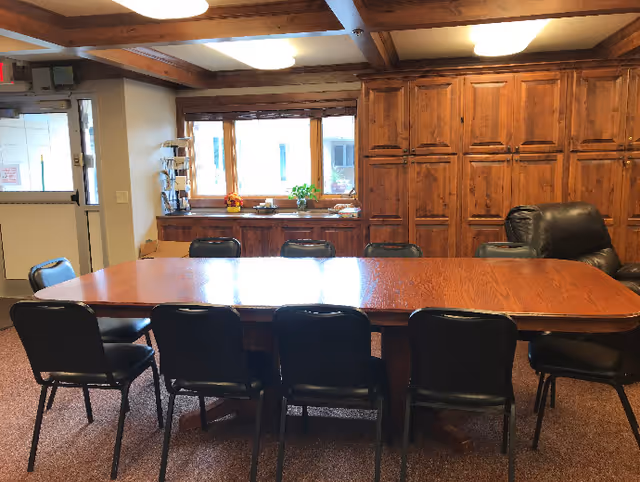 A meeting or dining room with a large wooden table surrounded by black chairs. The room has wooden cabinets along the back wall, a window with natural light, and a leather recliner chair to the right. The ceiling features wooden beams and there is a door to the left side of the room.