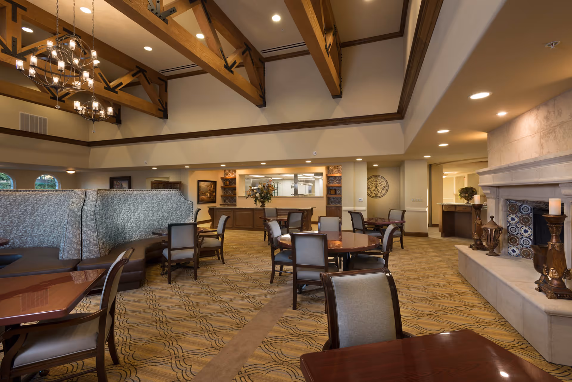 Spacious senior living dining room with wooden tables and chairs, patterned carpet, high ceiling with exposed wooden beams, chandeliers, and a large stone fireplace decorated with candles and ornamental tiles.