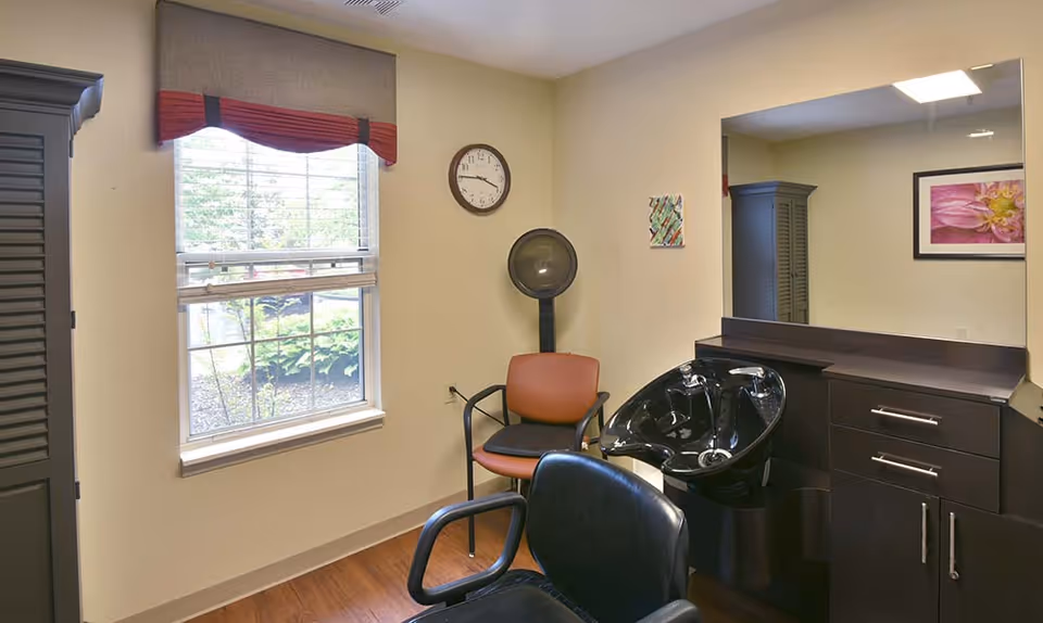 A small salon room with a shampoo basin, styling chairs, a hooded dryer, mirror, and a window looking outside.