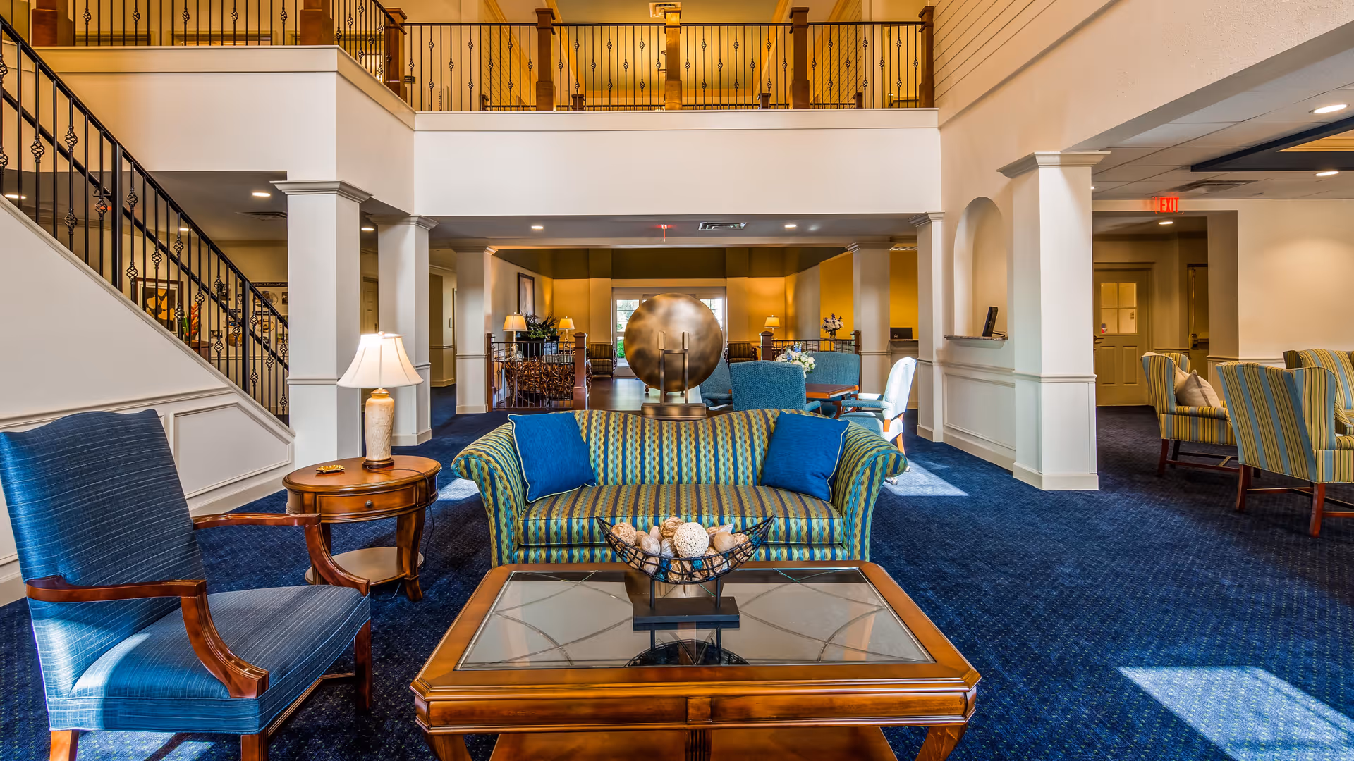 A spacious and well-lit living room area in a senior living facility featuring a blue and green striped sofa with two blue pillows, a wooden coffee table with a decorative bowl, a blue upholstered armchair, and a side table with a lamp. The room has blue carpet flooring, white walls with columns, and a staircase with black iron railings. The upper level balcony overlooks the seating area below.