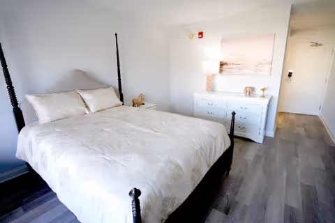 A bright bedroom with a large bed covered in a light-colored patterned bedspread, two pillows, a white dresser with a lamp and decorative items on top, a painting on the wall, and a door leading out of the room. The floor is wood with a grayish tone.