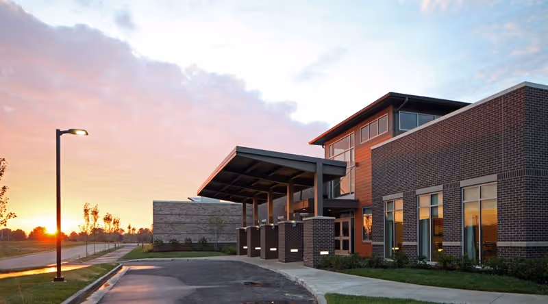 Exterior view of a modern senior living facility building at sunset with a covered entrance, brick and wood paneling, large windows, a street lamp, and a paved driveway.