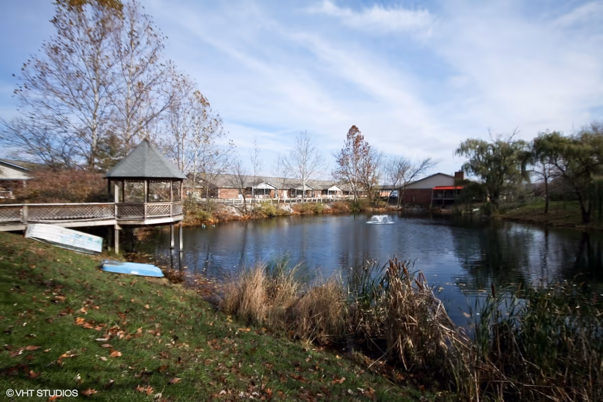 A peaceful outdoor scene featuring a small pond with a fountain in the center, surrounded by grass, trees with autumn leaves, and a wooden gazebo on the left side connected by a walkway. Residential buildings are visible in the background under a partly cloudy sky.