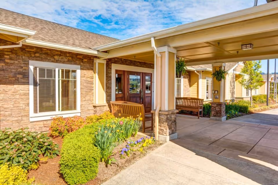 Entrance area of Arbor Oaks Terrace Memory Care Residence featuring a covered driveway with wooden benches, stone and beige siding exterior walls, a window with white blinds, and landscaped bushes and flowers under a partly cloudy sky.
