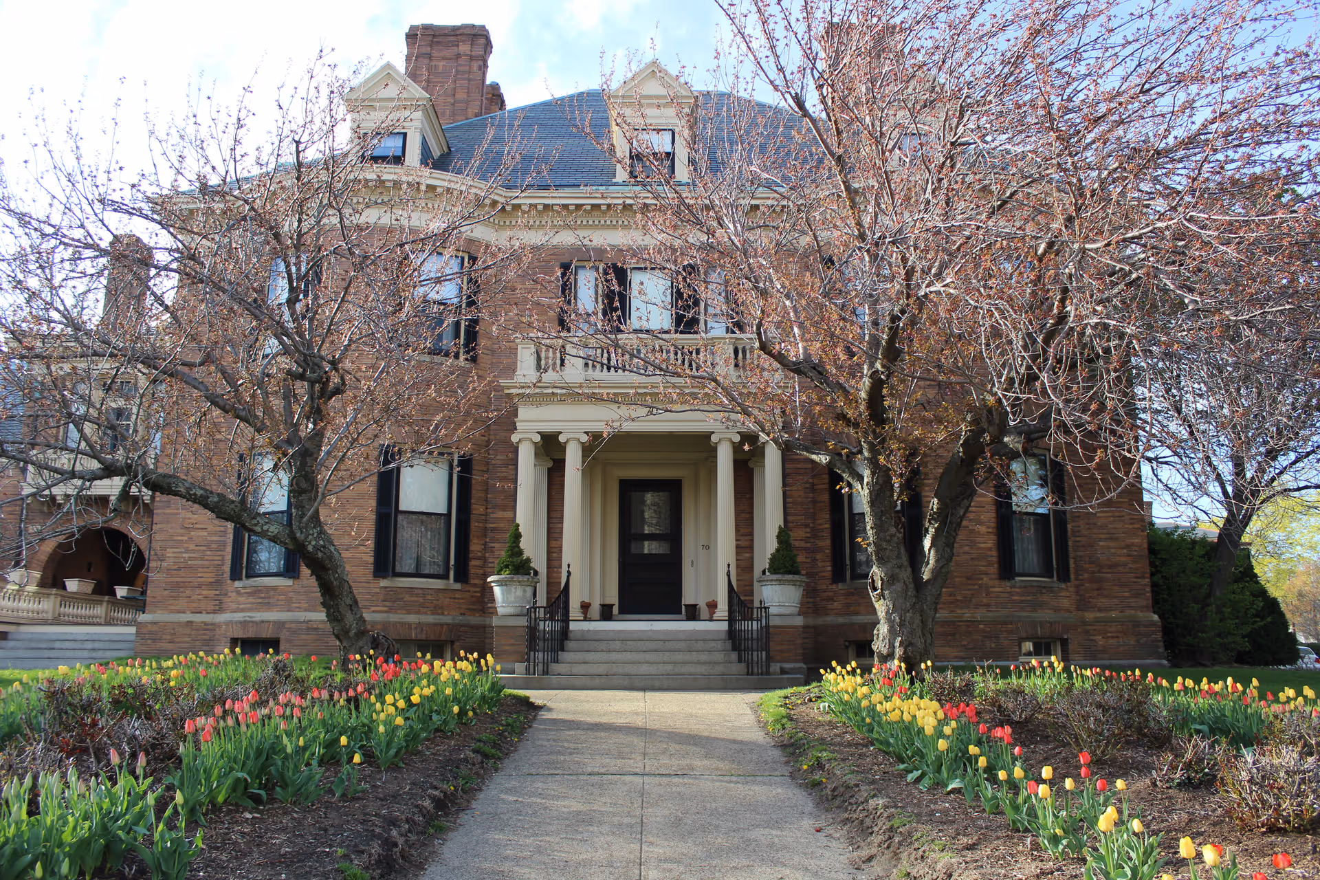 Front exterior view of a large, historic brick building with a central entrance featuring columns and a small balcony above. The building is surrounded by leafless trees and flower beds with red and yellow tulips along a concrete pathway leading to the entrance.