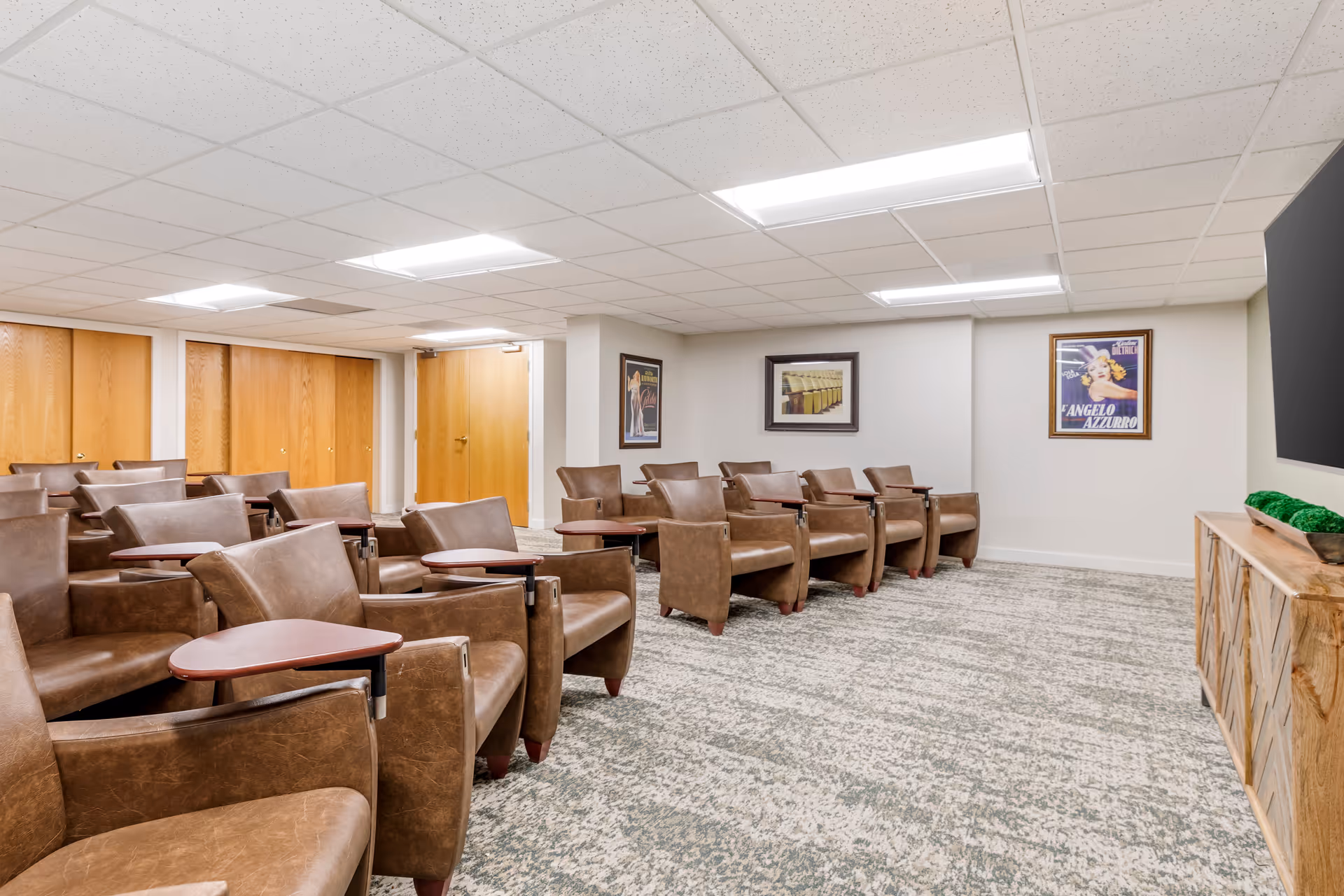 A small theater or media room with multiple rows of brown leather chairs equipped with wooden arm desks. The room has a carpeted floor, white walls with framed vintage posters, a wooden cabinet on the right side, and a large screen mounted on the wall.