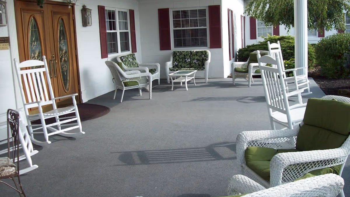 Outdoor covered porch area with white wicker chairs and sofas featuring green cushions, white rocking chairs, a small glass-top table, and a wooden double door entrance with decorative glass panels. The porch is adjacent to a white building with red shutters and windows, surrounded by greenery.
