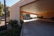 Covered driveway entrance to Mi Casa Nursing Center with a beige exterior wall and a landscaped area with bushes and a tree on the left side. Several parked cars are visible in the background under the covered area.