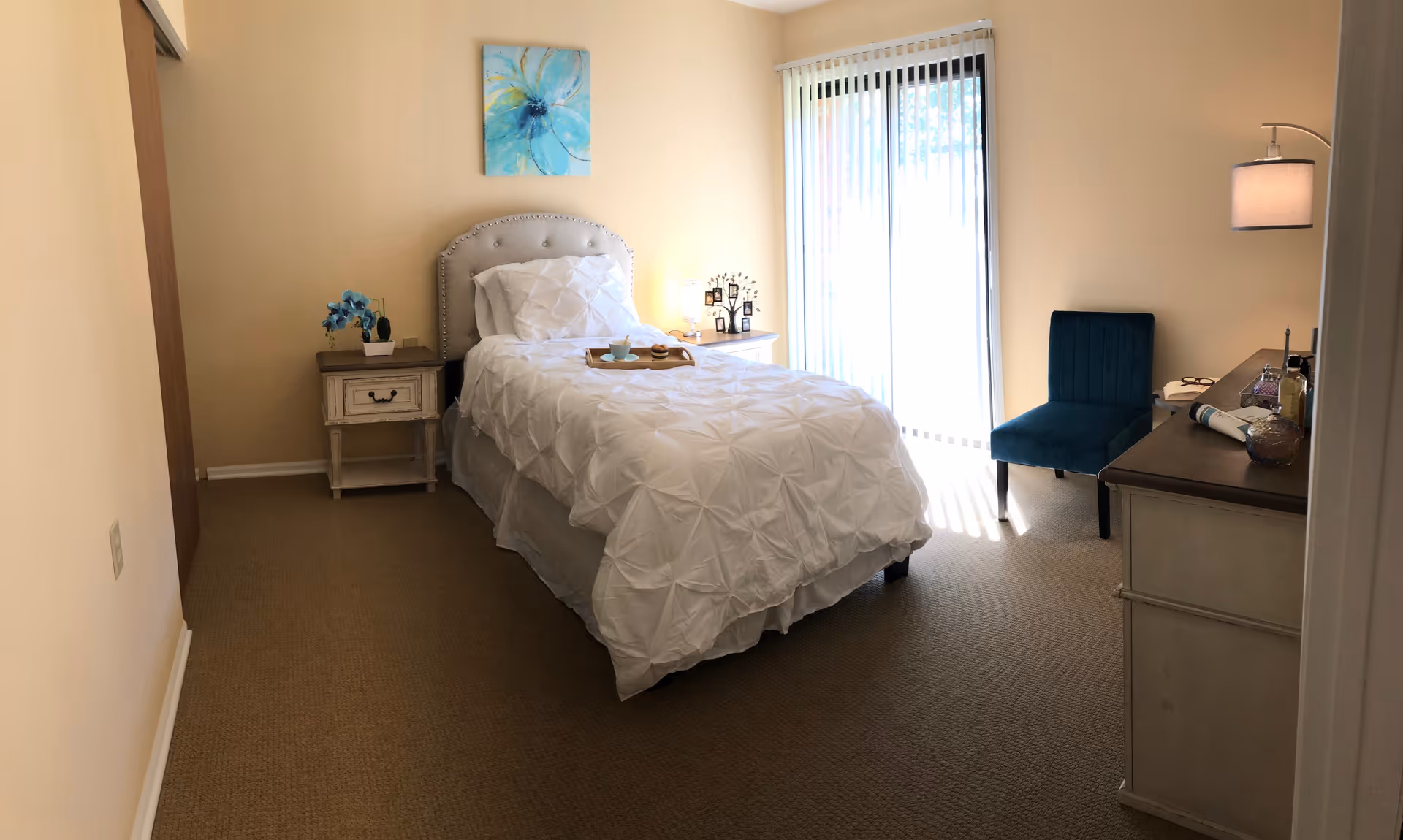 A neatly arranged bedroom with a single bed covered in a white comforter, a wooden nightstand with a blue orchid plant, a white dresser with various items on top, a blue upholstered chair, and a sliding glass door with vertical blinds letting in natural light. A blue floral painting hangs above the bed.