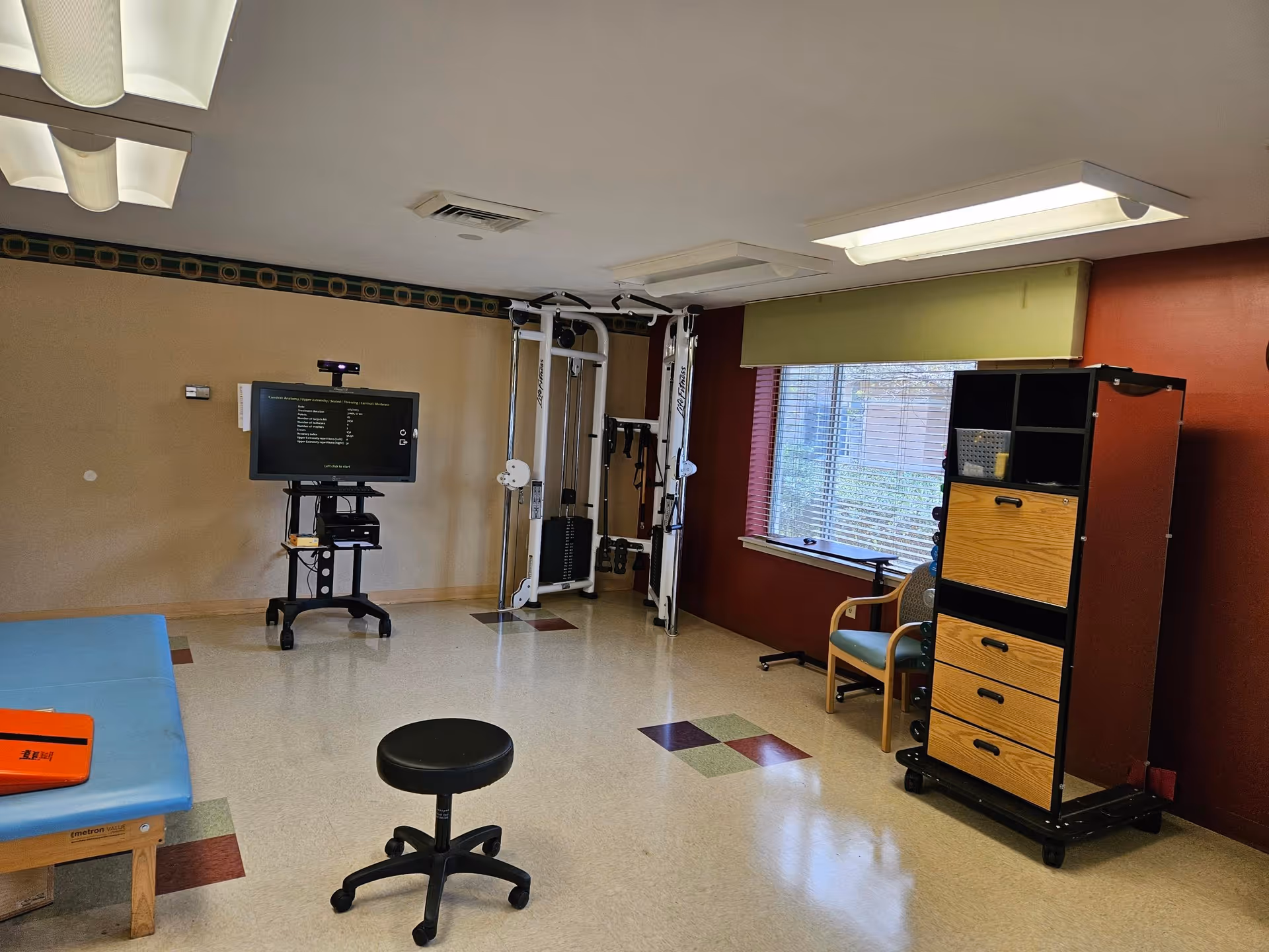 A therapy/exercise room with a TV on a cart, weight machine, treatment table, rolling stool, storage cabinet, and a window.