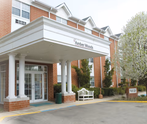 Entrance of Amber Woods At Harmar Village, showing a brick building with white columns supporting a covered entryway. There is a white bench near the entrance and a tree with white blossoms on the right side.