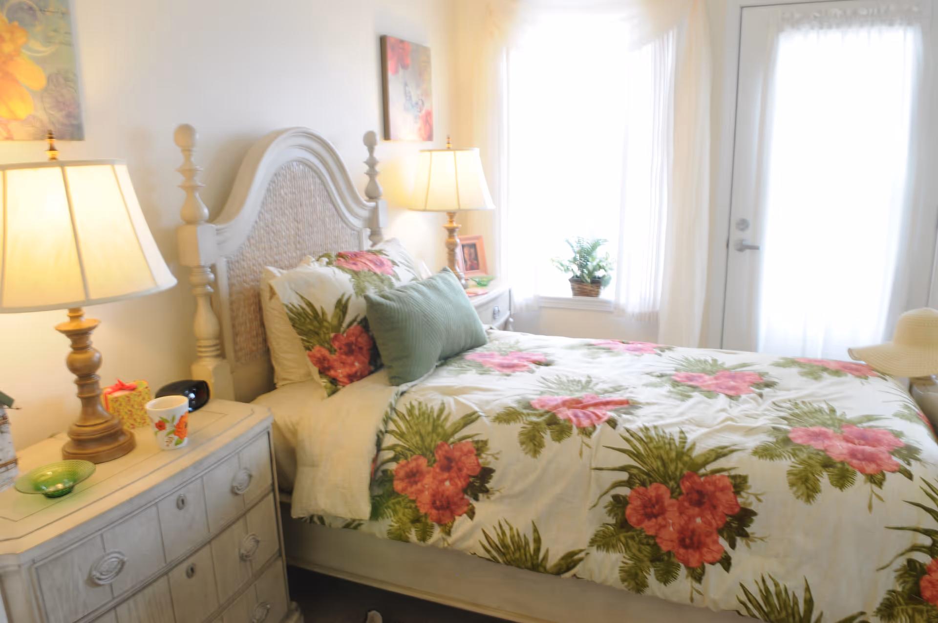 A bright bedroom with a bed featuring floral bedding with pink flowers and green leaves. There are two bedside tables with lamps on each side of the bed. A window with sheer white curtains and a door with similar curtains are visible in the background. A small potted plant sits on the windowsill.