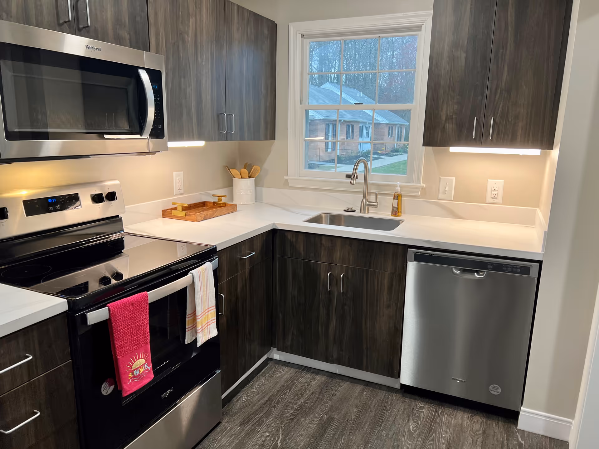Modern kitchen with dark wood cabinets, a stainless steel microwave, stove, and dishwasher. White countertops and a window above the sink showing a view of houses and trees outside. Two towels hang on the oven handle.