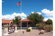 Front entrance of San Luis Care Center with a sign, an American flag on a flagpole, desert landscaping, and a single-story building under a blue sky.