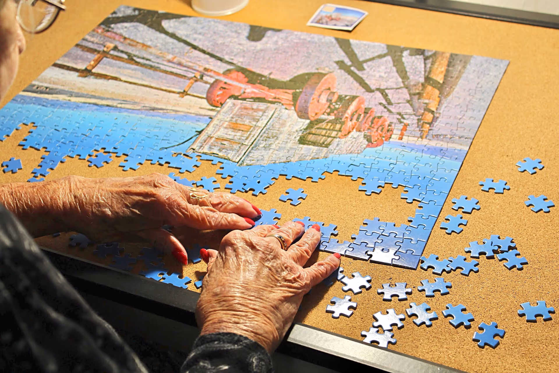 Close-up of an elderly person's hands working on assembling a jigsaw puzzle on a corkboard table. The puzzle depicts an outdoor scene with a red fire hydrant and a blue sky. Several puzzle pieces are scattered around the table.