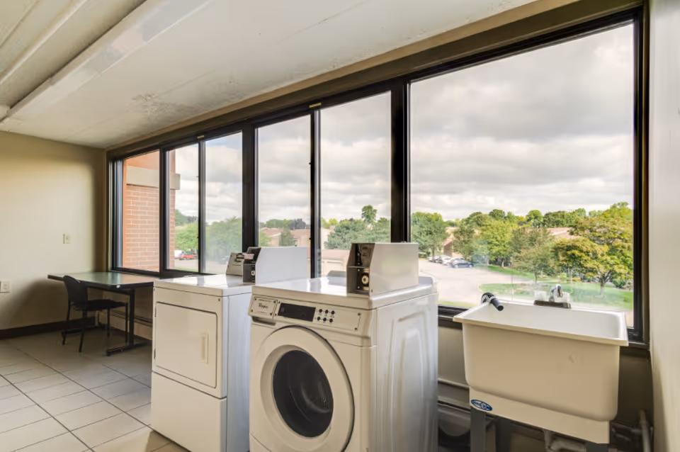 Laundry room with a washing machine and dryer next to a utility sink, large windows showing a view of trees and a parking lot outside, and a table with a chair against the wall.