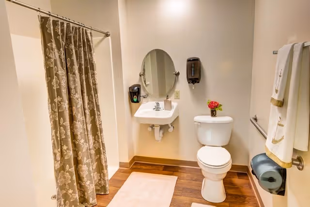 A clean bathroom with a floral-patterned shower curtain, a white sink with an oval mirror above it, a soap dispenser, a toilet with a small vase of flowers on the tank, a towel rack with white towels, and a toilet paper holder. The floor has wood-like tiles and a light-colored bath mat.