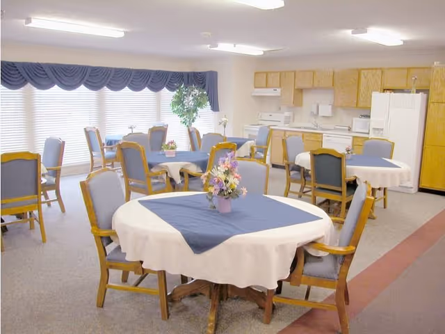 A dining area in a senior living facility with multiple round tables covered with white tablecloths and blue table runners. Each table has a small floral centerpiece. The room has several wooden chairs with blue cushions, large windows with blue valances, and a kitchen area with wooden cabinets, a refrigerator, stove, and microwave in the background.
