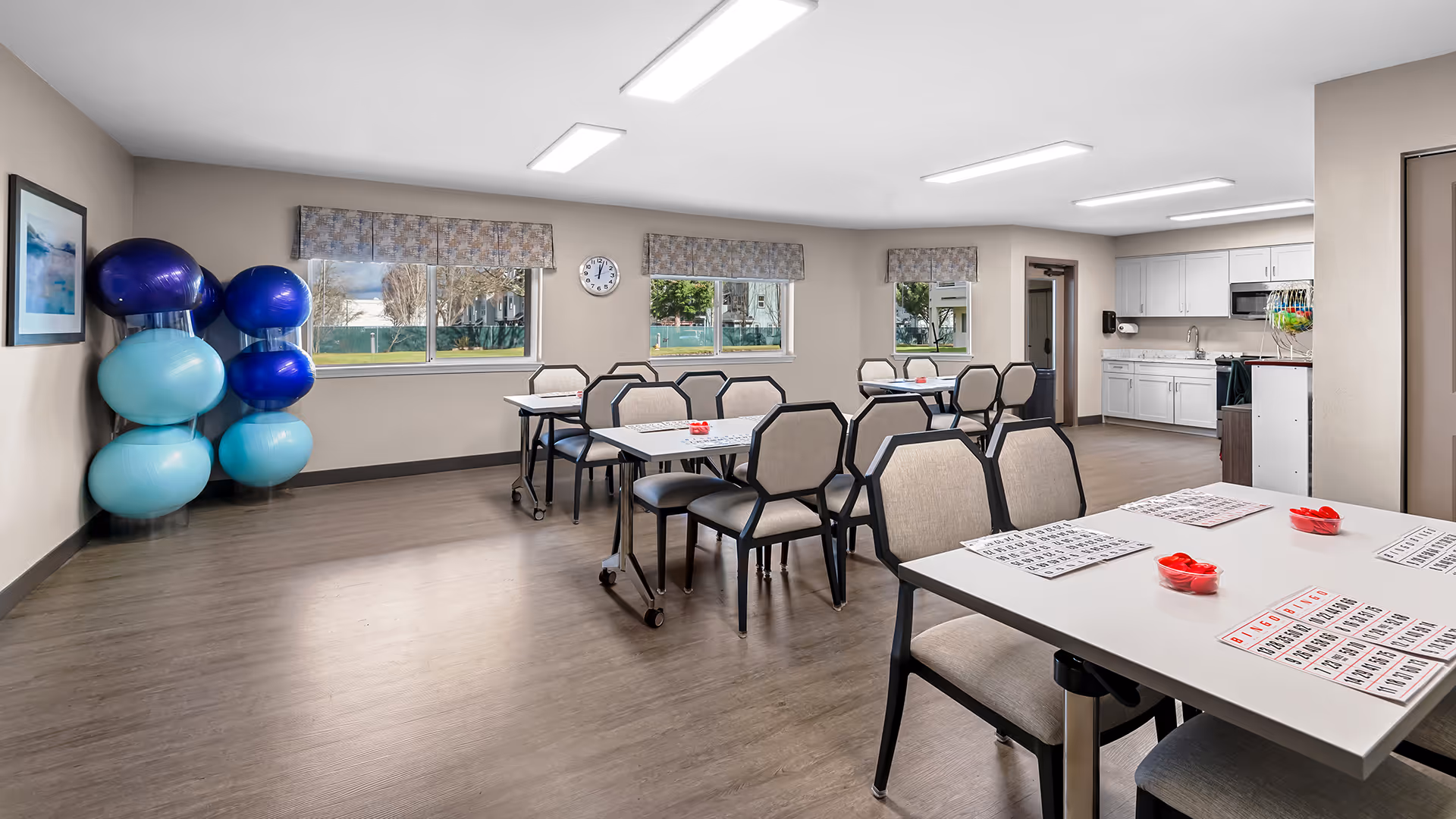 A bright activity room with several tables and chairs arranged for a bingo game, with bingo cards and red bingo chips on the tables. The room has large windows with patterned valances, a wall clock, and a rack holding various exercise balls in shades of blue. There is a kitchenette area with white cabinets, a sink, and a microwave in the background.