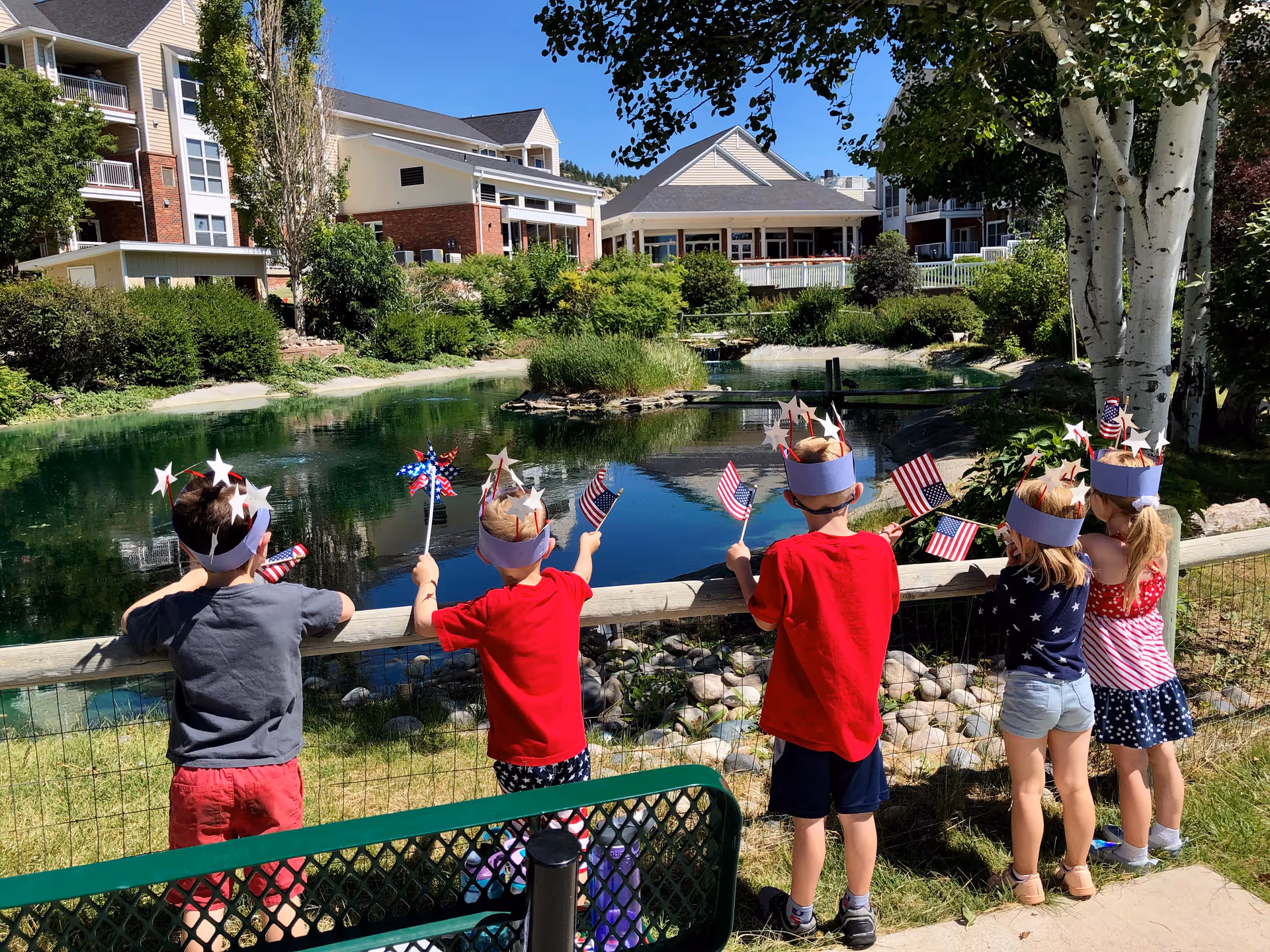 Five children wearing patriotic headbands and holding small American flags stand by a fence overlooking a pond with greenery and buildings in the background on a sunny day.
