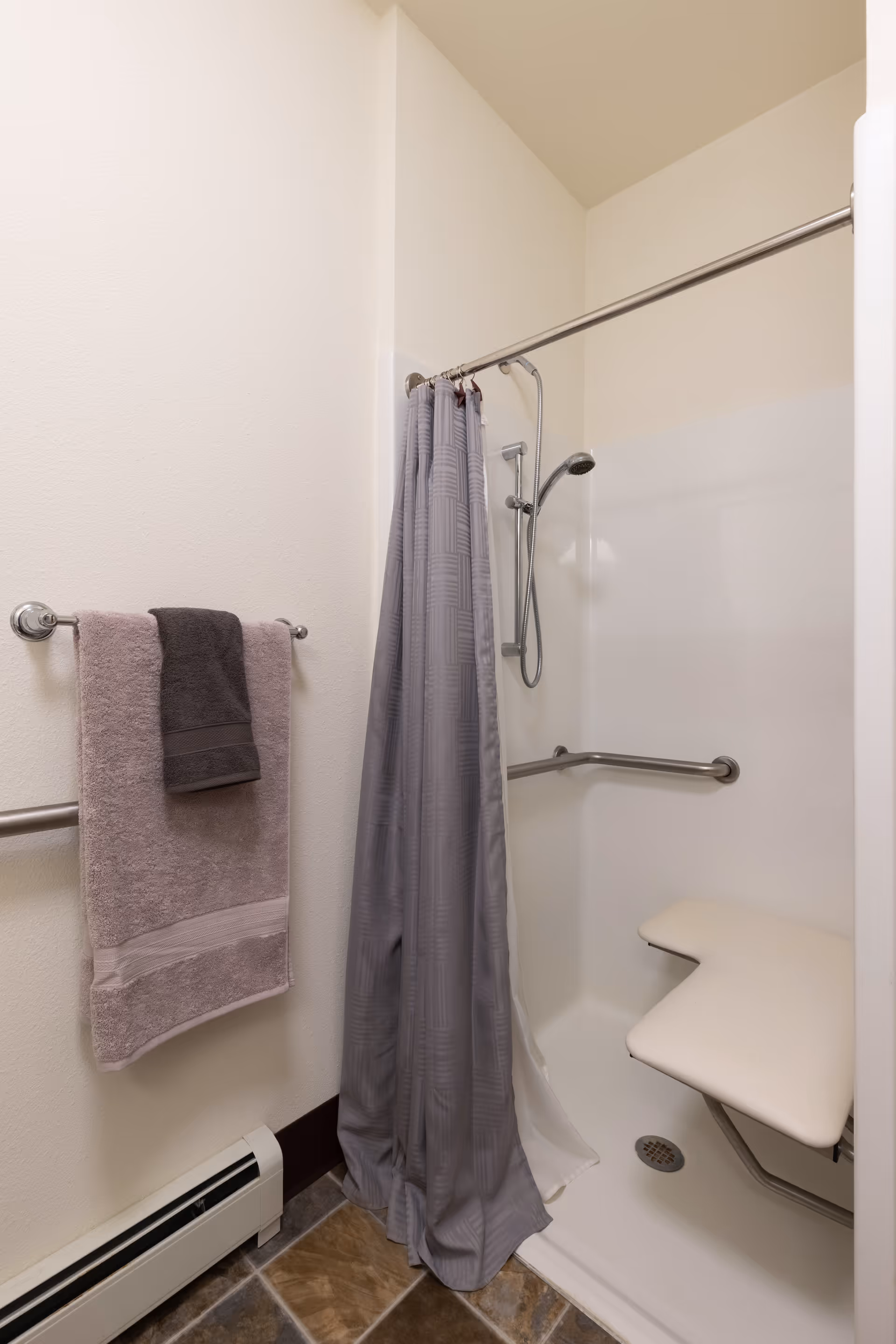 A bathroom shower area with a gray shower curtain, a handheld showerhead, a fold-down shower seat, and grab bars. A towel rack on the wall holds two towels, one light purple and one dark gray. The floor has brown and gray tiles.