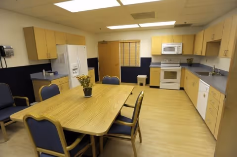 A kitchen and dining area with light wood cabinets, a white refrigerator, microwave, stove, and dishwasher. There is a wooden dining table with six blue cushioned chairs around it. The room has a light wood floor and fluorescent ceiling lights.