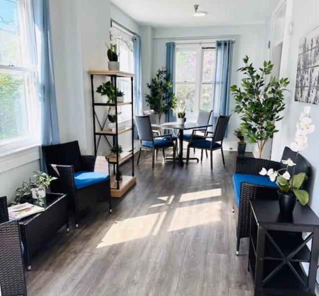 A bright and airy sitting area in a senior living community with large windows letting in natural light. The room features blue cushioned wicker chairs, a small black side table with a white orchid plant, a black metal and wood shelving unit with potted plants, and a round table with four chairs near the window. Light blue curtains frame the windows, and there are several green plants adding a touch of nature to the space. The floor is a light wood laminate.