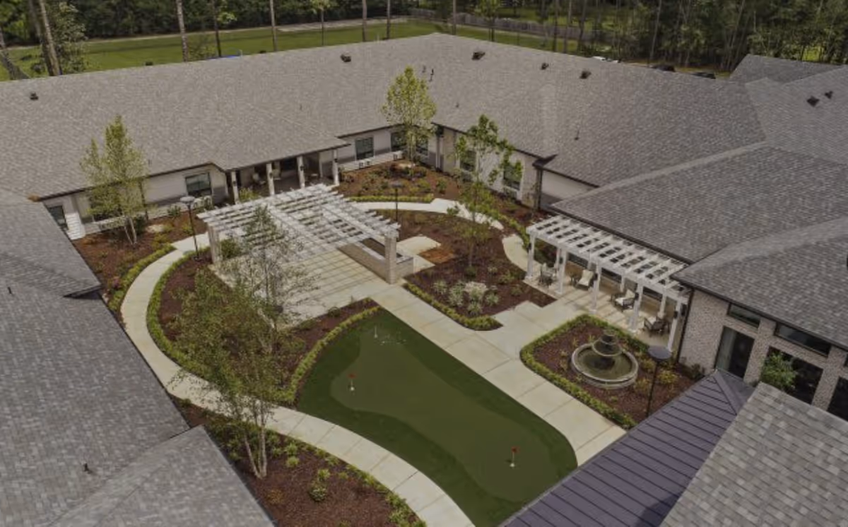 Aerial view of a senior living facility courtyard featuring a putting green, walking paths, landscaped garden beds, trees, a water fountain, and shaded seating areas with pergolas surrounded by the building's roof.