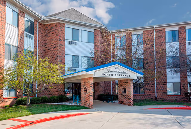 Exterior view of Brenden Gardens senior living facility showing the north entrance with a covered driveway, brick and white siding building, trees, and a clear sky.