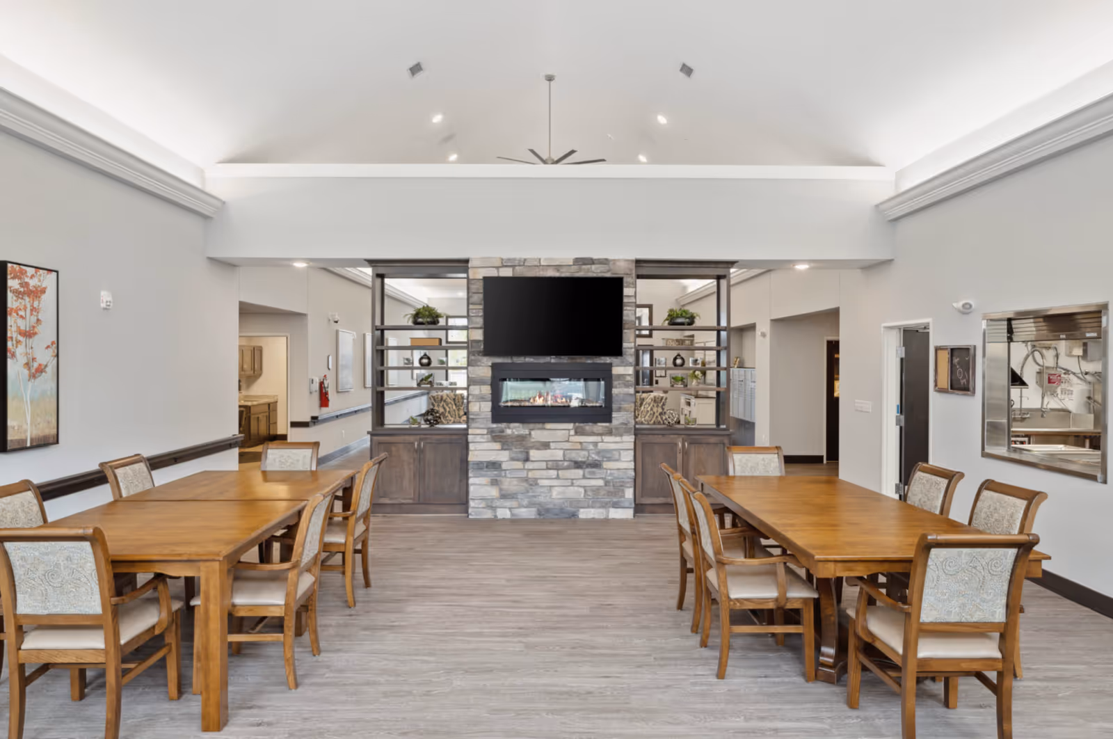 Bright communal dining room with wooden tables and chairs facing a stone fireplace and wall-mounted TV under a vaulted ceiling.