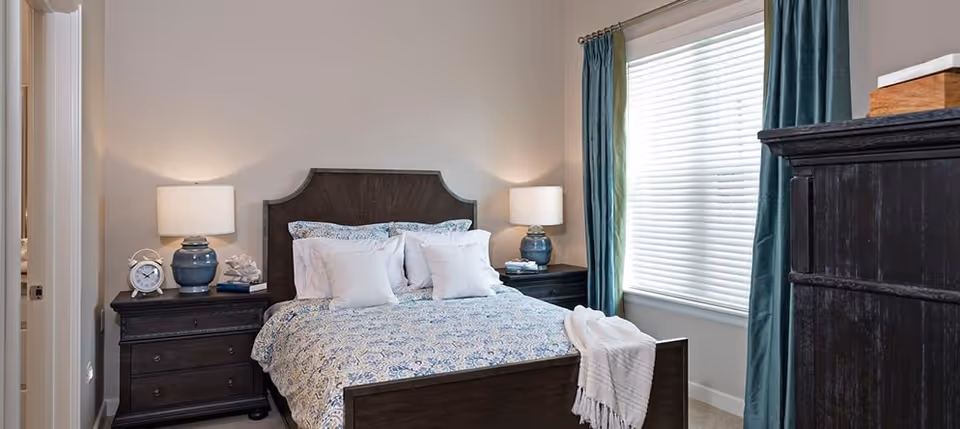 A cozy bedroom featuring a dark wooden bed with a blue and white patterned bedspread and multiple white pillows. There are matching dark wooden nightstands on each side of the bed, each with a blue lamp and decorative items. A window with white blinds and teal curtains is on the right side, and a dark wooden dresser is partially visible on the right edge of the image.