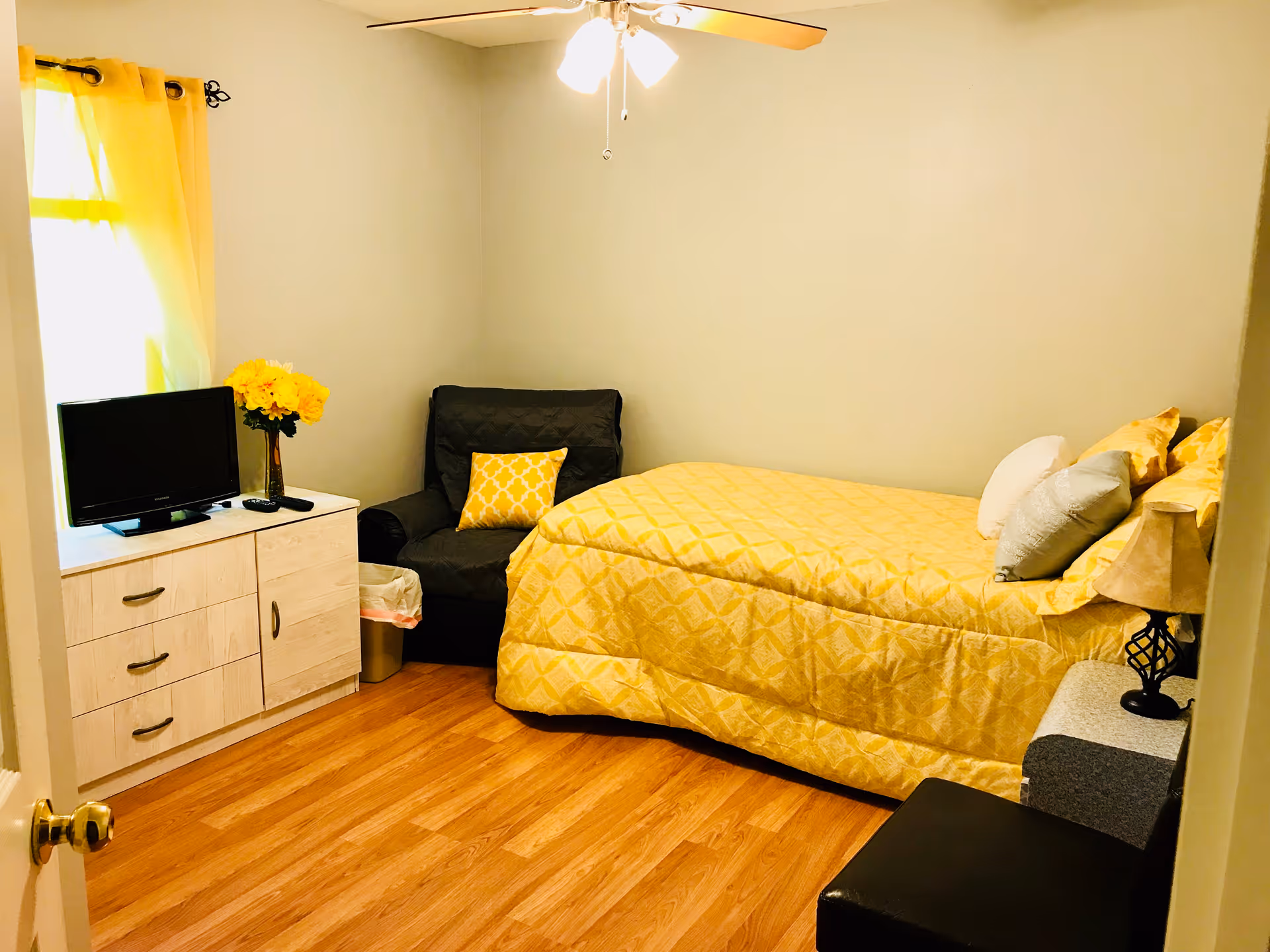 A cozy bedroom with a single bed covered in a yellow patterned comforter and multiple pillows. There is a black armchair with a yellow cushion next to the bed. A white dresser with drawers holds a small TV and a vase of yellow flowers. The room has wooden flooring, light-colored walls, a ceiling fan with lights, and a window with yellow curtains letting in natural light.