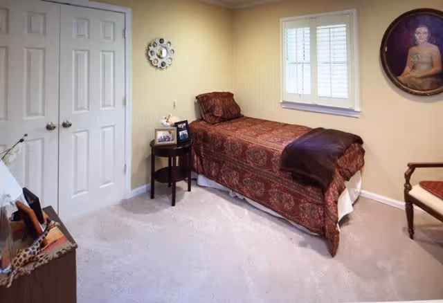 A small bedroom with a single bed covered in a patterned red bedspread and a dark brown throw blanket. Next to the bed is a round wooden side table with framed photos and a small decorative item. The room has beige walls, a window with white shutters, a double-door closet, a wooden chair with a red cushion, and a framed portrait hanging on the wall.
