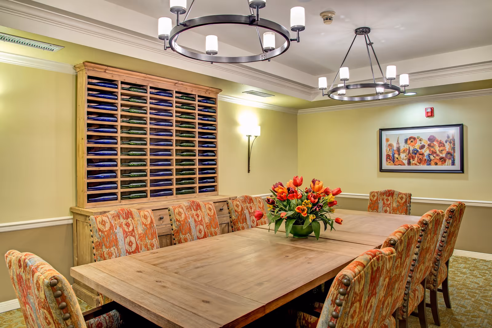 A well-lit dining room with a large wooden table surrounded by upholstered chairs with a red and orange pattern. A vase with red and orange tulips is placed in the center of the table. On the wall behind the table is a wooden cabinet with multiple compartments holding folded blankets or linens. The walls are painted light yellow with white trim, and there is a framed floral artwork on one wall. Two modern circular chandeliers hang from the ceiling.