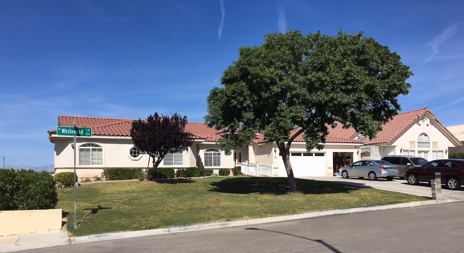 Exterior view of a single-story residential building with a red tile roof, beige walls, and several windows. There is a green lawn with two trees in front of the building, and three cars parked in the driveway. A street sign labeled 'N Whitewind Ln 100' is visible on the left side of the image under a clear blue sky.