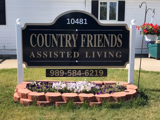 A black and gold sign for Country Friends Assisted Living with the address number 10481 and phone number 989-584-6219, mounted on white posts surrounded by a small flower bed with purple and white flowers in front of a white building with black shutters.