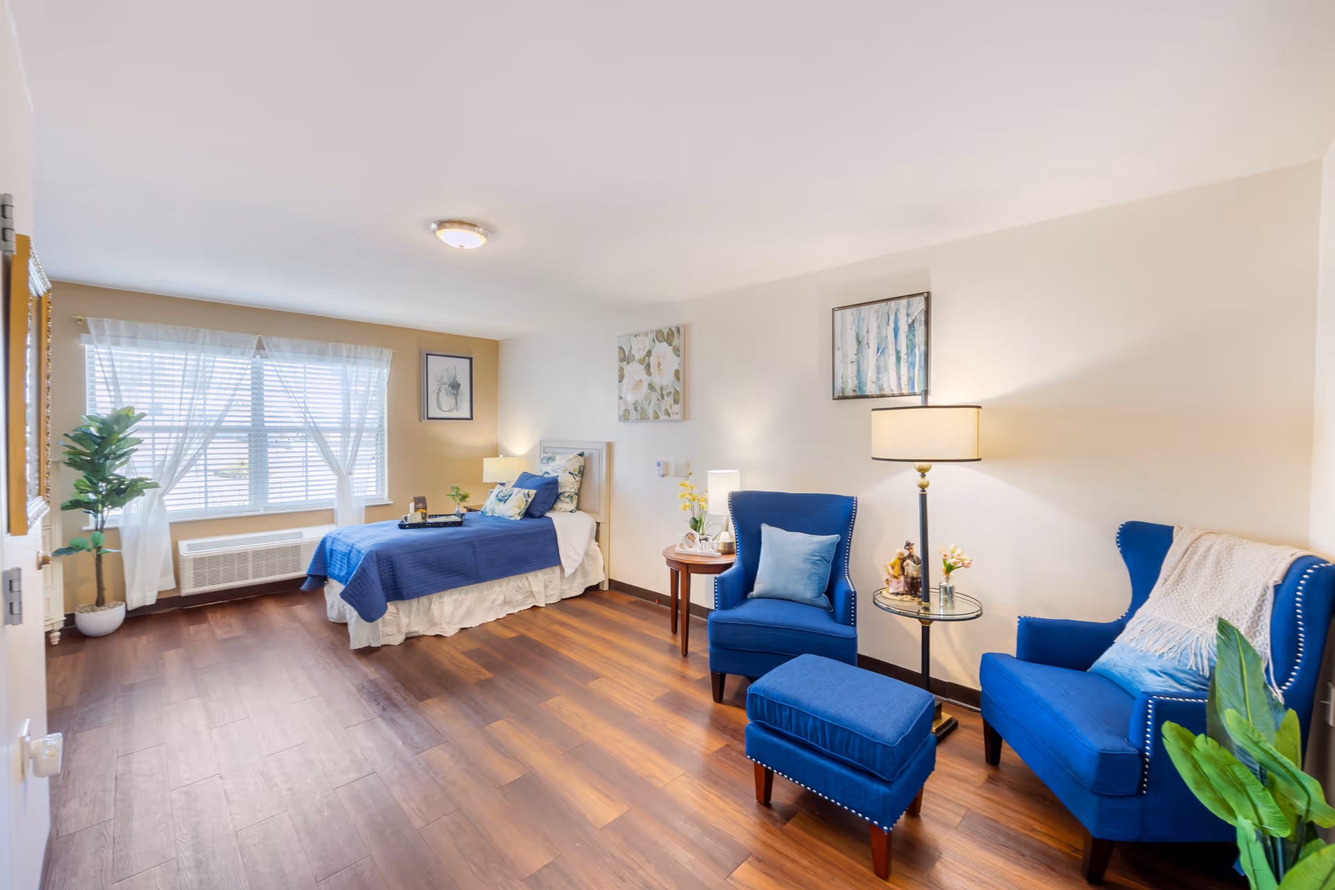 A bright and spacious senior living bedroom with a single bed covered in blue bedding, two blue armchairs with a matching ottoman, a floor lamp, side tables with decorative items, and large windows with sheer white curtains letting in natural light. The room has wooden flooring and light-colored walls adorned with framed artwork.