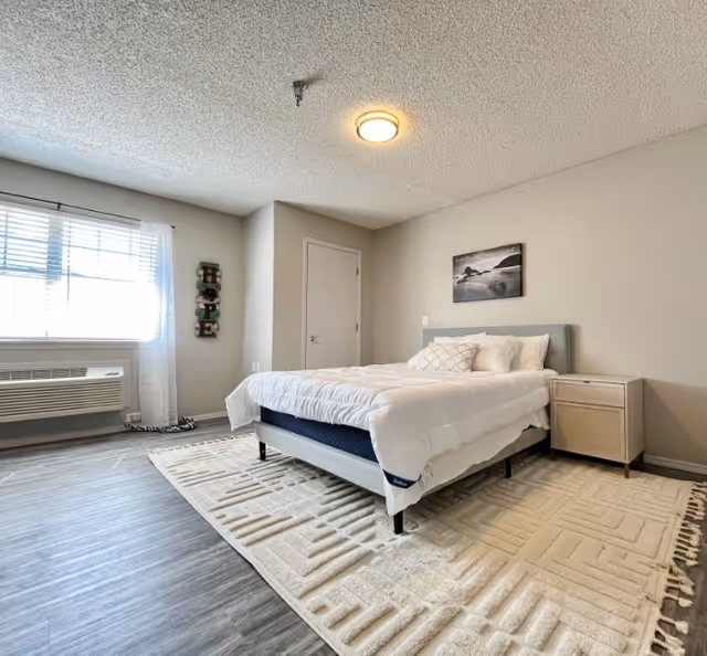 A tidy bedroom in a senior living facility with a bed, rug, and window.