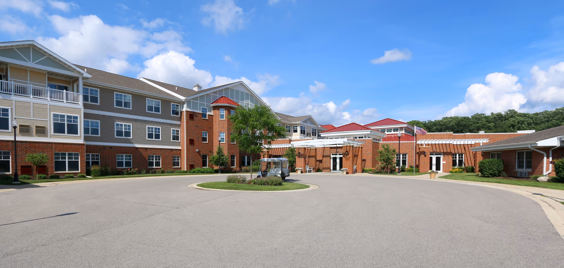 Exterior view of a senior living facility with multiple connected buildings featuring brick and beige siding, a circular driveway with a small landscaped island in the center, and a clear blue sky with some clouds.