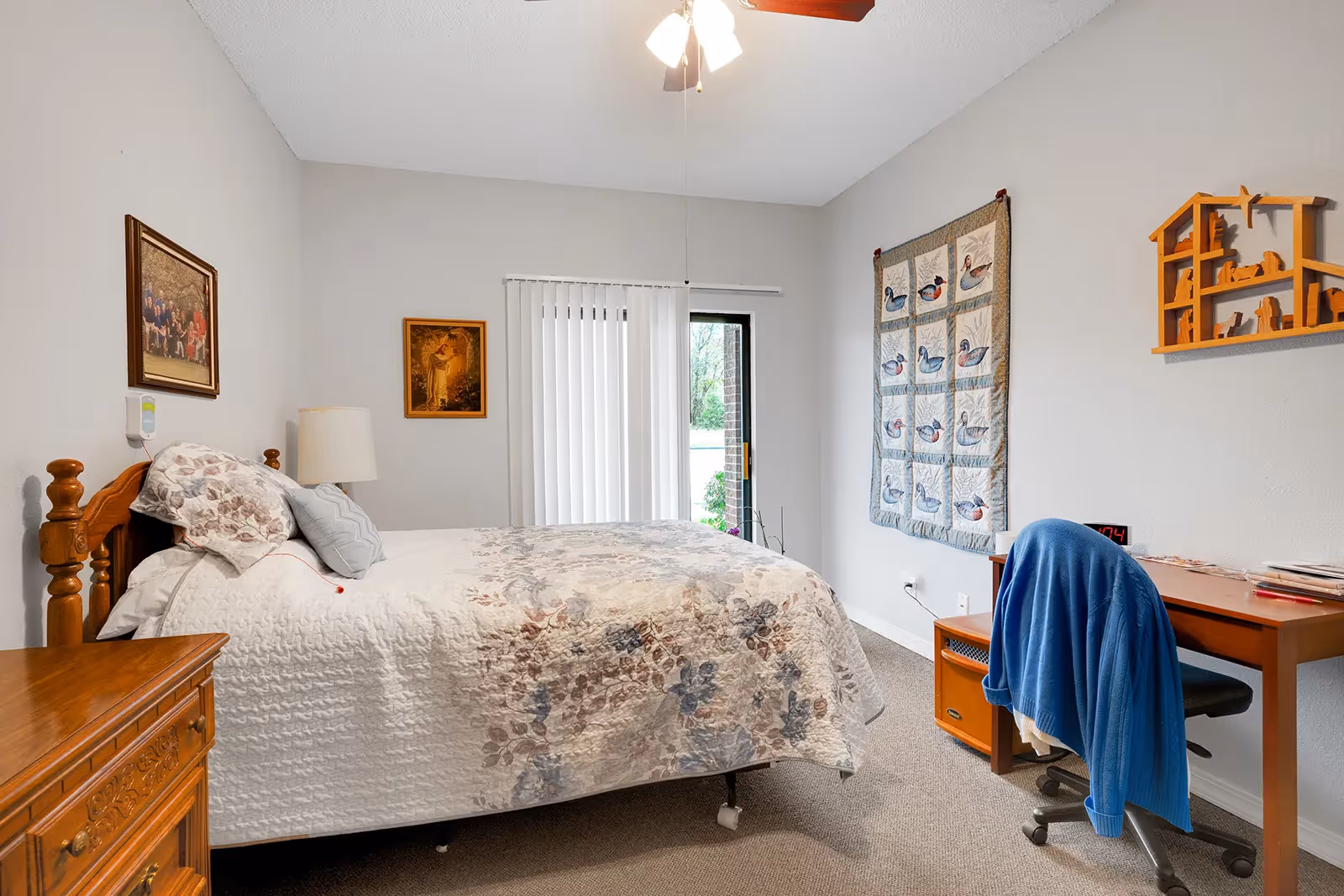 A cozy bedroom with a wooden bed frame and floral bedding. There is a wooden nightstand with a lamp on the left side of the bed. On the right side of the room, there is a wooden desk with a blue jacket draped over a black office chair. The wall above the desk has a decorative quilt with duck patterns and a wooden wall shelf shaped like a house with animal figurines. Two framed pictures hang on the wall above the bed. Vertical blinds cover a sliding glass door that leads outside.