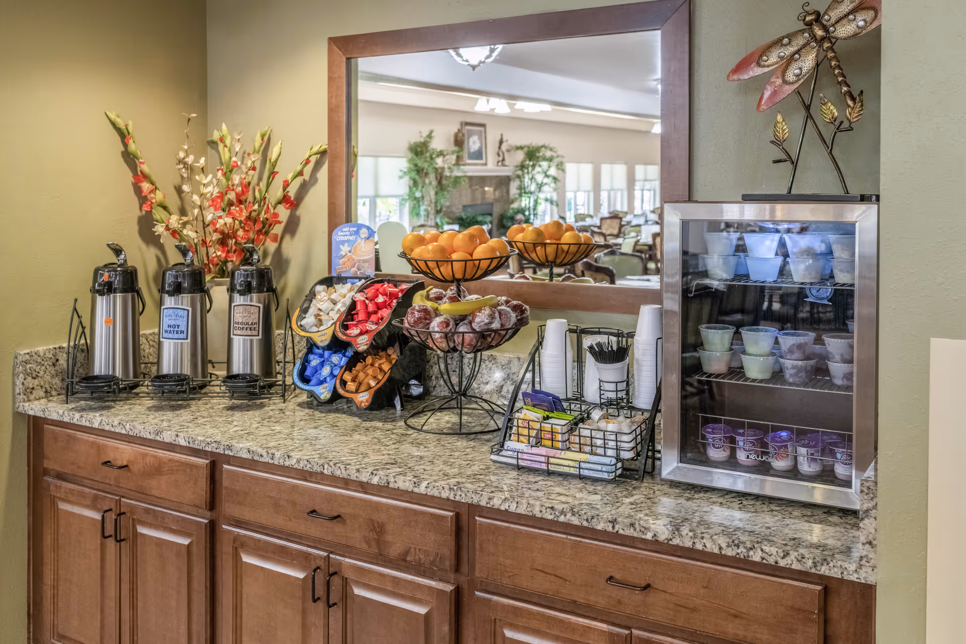 A countertop with three beverage dispensers labeled hot water, regular coffee, and decaf coffee, a tiered fruit basket with oranges and apples, a rack with various creamers and sweeteners, disposable cups, and a small refrigerator containing yogurt cups. A large mirror reflects a seating area with chairs and plants in the background.