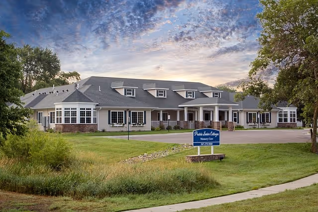 Exterior view of Prairie Senior Cottages of Albert Lea, a single-story building with multiple dormer windows, a covered entrance, and surrounded by green grass and trees under a partly cloudy sky at sunset.