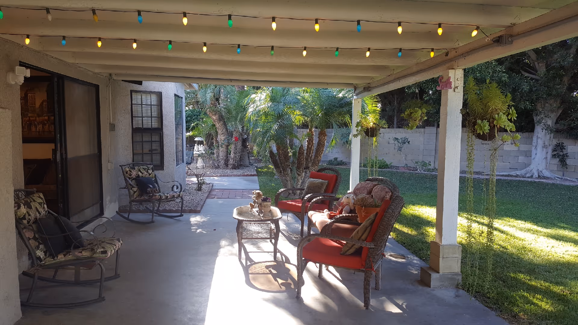 Covered patio area with outdoor seating including cushioned chairs and a small table. The patio is decorated with hanging plants and string lights with colorful bulbs. The area opens to a garden with palm trees and a grassy lawn enclosed by a brick wall.
