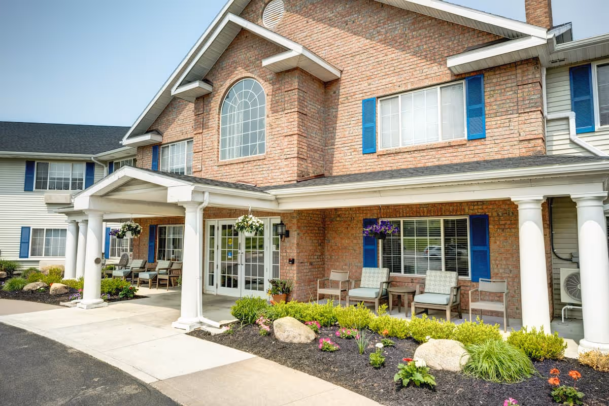 Front exterior view of a senior living facility named Evergreen Place featuring a brick facade with white columns supporting a covered entrance. There are several chairs and hanging flower baskets on the porch, with landscaped flower beds and shrubs in front.