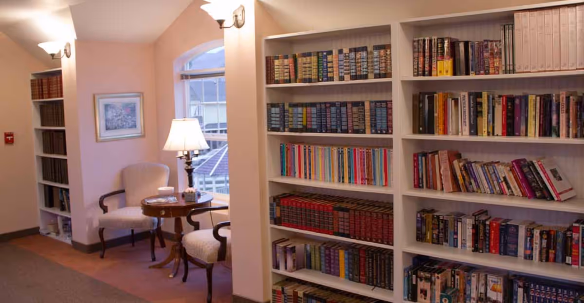 A cozy reading nook in a retirement facility featuring two upholstered armchairs around a small wooden table with a lamp. The space is softly lit with wall sconces and natural light from a window. There are built-in bookshelves filled with books on the right and left sides of the area.