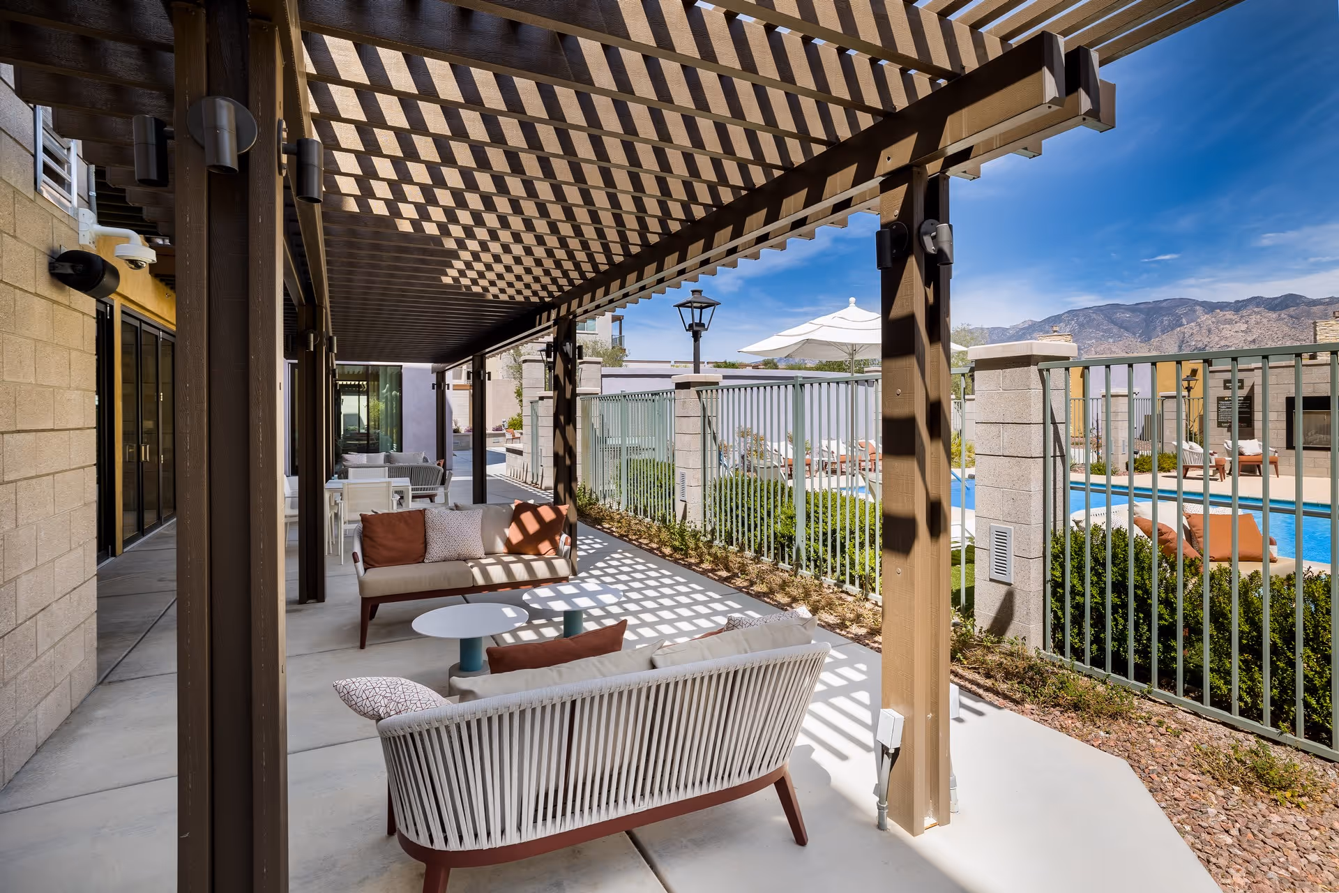 Outdoor patio area with cushioned seating and small tables under a wooden pergola casting patterned shadows. Beyond the fenced area is a swimming pool with lounge chairs and umbrellas, set against a backdrop of mountains and a clear blue sky.